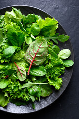 A mix of fresh, raw salad greens on a black plate. Includes lettuce, escarole, spinach, romaine, radicchio, lamb's lettuce, arugula. Healthy, low-calorie food ideal for a diet. Top view