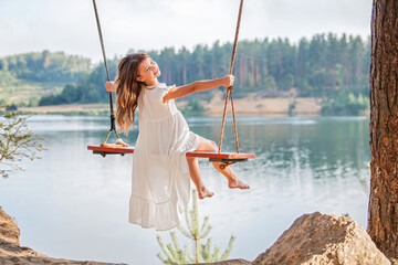 A girl in the forest swinging on a swing. Rope swing on a forest lake. Barefoot girl in a white dress with long hair.