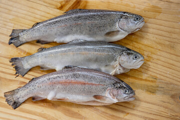 Carving three raw fresh trouts on the old wooden cutting board. Cooking whole freshwater rainbow forel. Oncorhynchus mykiss on the chopping plate. Wood background. Close up. Copy space. Top view
