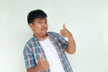 Excited and happy Asian man gives thumbs up hand gesture of approval, isolated by gray background.