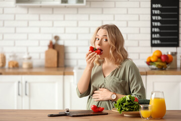 Young pregnant woman eating bell pepper at table in kitchen