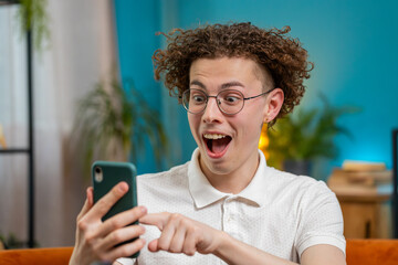 Surprised excited Caucasian young man winner holding smartphone reading good news amazed by online...