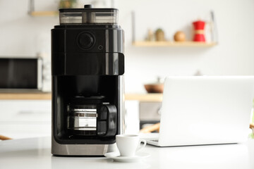 Modern coffee machine with cup and laptop on table in kitchen
