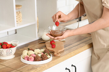 Young woman dipping fresh strawberry into bowl with melted chocolate in kitchen