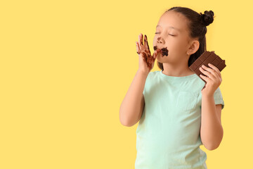 Funny little Asian girl with chocolate licking her finger on yellow background