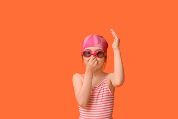Cute little girl in swimsuit with swimming hat and goggles holding her nose on orange background