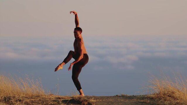 Male ballet dancer doing a choreographic dance move standing on a rock at sunset. Heavenly clouds are in the background