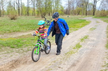 Naklejka premium Father running by child learning cycling on footpath at park, teaching kid to ride bicycle on road