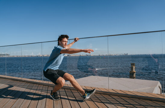 Man performing a single-leg squat outdoors by the waterfront. He is wearing a blue t-shirt and black shorts. Clear blue sky and cityscape in the background, emphasizing fitness and balance - Powered by Adobe