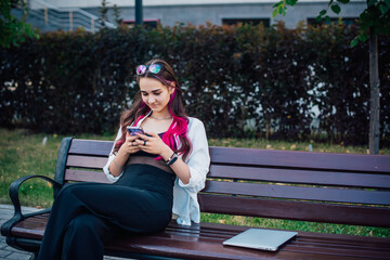 Fototapeta premium Portrait shot of a young brunette woman messaging on mobile phone while sitting on the bench outdoors. Beautiful female person using smartphone outside.