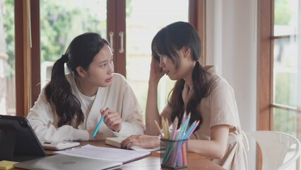 Two Asian university female friends are tutoring their friend who doesn't understand by discussing the course content and explaining through books and study materials at their home