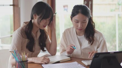 Two Asian university female friends are tutoring their friend who doesn't understand by discussing the course content and explaining through books and study materials at their home