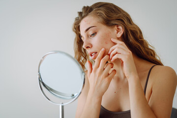 A beautiful woman looking in the mirror, touching her face with her hand, examines acne,...