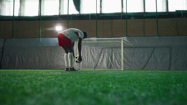Black soccer player exercising penalty in training hall, scoring goal, back view. Sport training of national team before match and competition, African American man is forward or halfback, empty gate