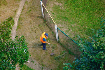 Municipal worker in overalls collecting mown grass with rake after mowing, viewed from above, maintaining a public lawn. Top view of unrecognizable municipal worker raking grass after mowing