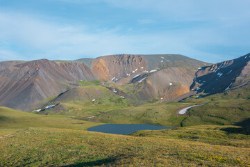 Fototapeta premium Alpine small lake in sunlit green valley among grassy hills against colorful mountain range under cloudy sky. Beautiful lake among high mountains of many vivid colors in sunlight in changeable weather