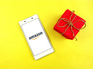 Closeup amazon com app on white phone screen & red gift box, gold ribbon bow. Amazon logo of online...