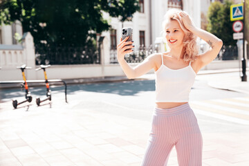 Young beautiful smiling blond hipster woman in trendy summer clothes. Carefree female posing in the street at sunny day. Positive model outdoors at sunset. Cheerful and happy. Takes selfie