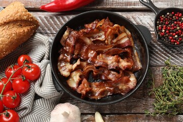 Delicious bacon slices in frying pan and products on wooden table, flat lay