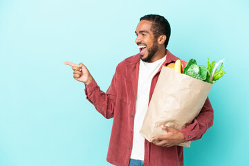 Young latin man holding a grocery shopping bag isolated on blue background pointing finger to the...