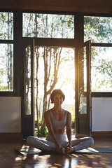 Young happy fit sporty slim Hispanic woman yoga coach, stretching sport trainer wearing grey top and pants sitting on mat in modern yoga gym studio in nature park looking at camera. Vertical portrait.