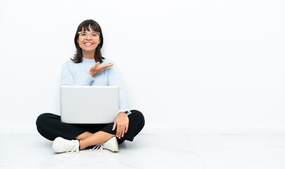 Young mixed race woman sitting on the floor with laptop isolated on white background presenting an idea while looking smiling towards