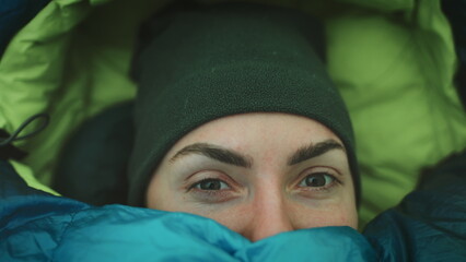 Person wake up in camp tent sleeping bag. Close up portrait of tourist girl opening eyes in cold morning. Female travel wilderness vacation, tourism and camping in outdoor trip. Slow motion 4K