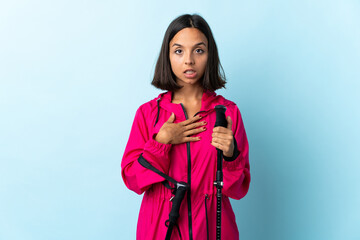 Young latin woman with backpack and trekking poles isolated on blue background surprised and shocked while looking right