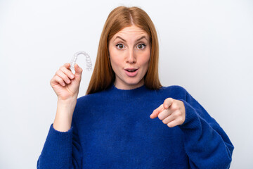 Young redhead woman holding invisible braces isolated on white background surprised and pointing...