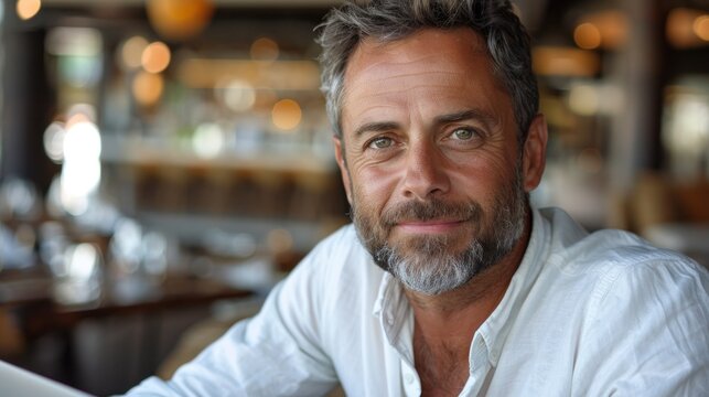 A middle-aged man with graying hair and a beard smiles at the camera in a restaurant setting