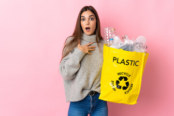 Young caucasian woman holding a bag full of plastic bottles to recycle isolated on pink background surprised and shocked while looking right