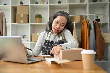 Senior female small business owner confirming the order on phone and using laptop at her shop