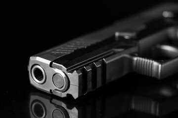 Close-up of the muzzle and barrel of a semi-automatic pistol with a metal surface. The weapon sits sideways against a reflective black surface, providing a detailed view of the front end.