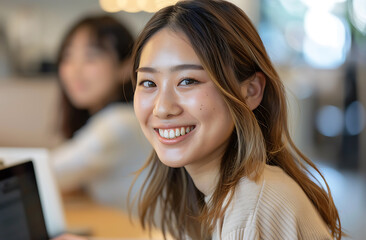 Obraz premium Japanese woman her early mid20s smiling while sitting office desk holding looking into camera one hand top of laptop keyboard She shoulderlength hair wear lightcolored casual attire blurred