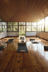 Modern studio for yoga meditation practices background with mats on wooden floor. Empty clean gym room interior with big windows for group retreats sessions in natural forest garden area. Vertical