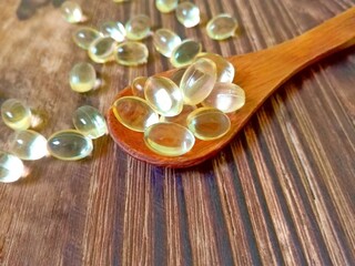 Vitamin D yellow pills on wood spoon on wooden background & soft sun light. Medicine gel tablets of vitamin d on table. Yellow vitamin D oil capsule keeps healthy while lack of sun - medicine concept