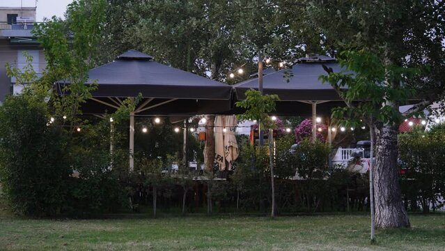 Outdoor terrace of resort town restaurant with dark umbrellas and glowing garland of retro light bulbs in evening. Cozy green area for relaxing and eating restaurant food without people.