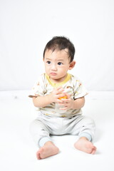 A cute baby with a sad expression, sitting and holding a milk bottle filled with water. The baby wears a light-colored outfit with animal prints, seated on a white background.