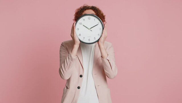 Happy smiling Caucasian curly haired man in jacket holding office clock hiding behind, checking time on watch, running late to work, being in delay, deadline obscuring face on pink background indoors