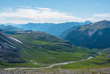 Aerial top view to sunlit green alpine valley with creek among big hills and large rocky mountains silhouettes in bright sun under clouds in blue sky. High snowy mountain range far away in sunny day.