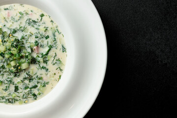 Cold soup with fresh cucumbers, sausage, broth and sour cream in a plate-hat on a black background. A traditional dish is okroshka