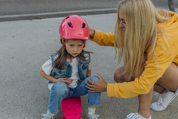 Mom helping her daughter to put on a helmet and ride a skateboard