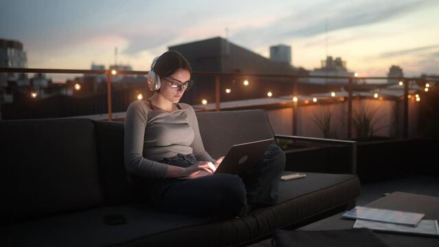 Woman working on laptop outdoors at twilight in a modern city setting, a focused freelancer. A woman with headphones is sitting on a rooftop sofa, working on her laptop as the sunset