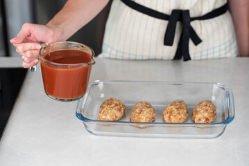Female chef in an apron preparing meat or cutlets patties with rice, baking them in a dish with tomato sauce. Home cooking, traditional recipes, and culinary preparation.