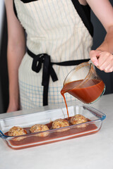 Female chef in an apron preparing meat or cutlets patties with rice, baking them in a dish with tomato sauce. Home cooking, traditional recipes, and culinary preparation.