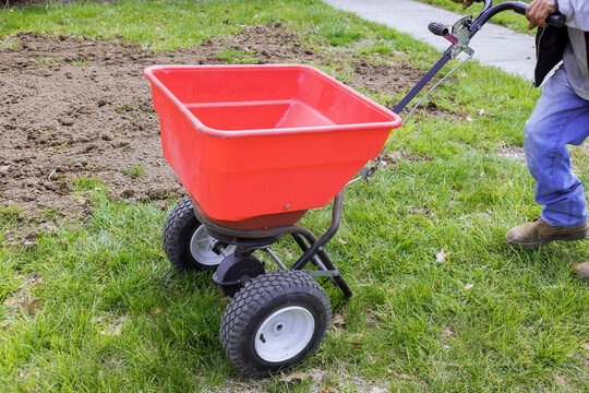 An individual sowing lawn grass seeds with drop lawn spreader in residential backyard