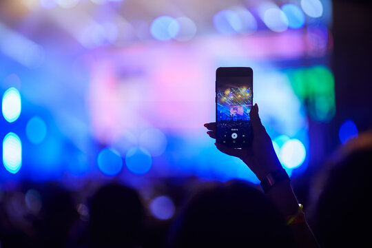 Concertgoer films live music event with smartphone, hand raised above crowd, capturing stage lights, performers in blurred background. Energetic festival atmosphere, fans enjoy night performance.