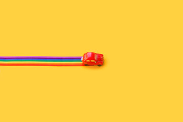 Blue toy car, rainbow ribbon flag trail road, yellow background. June gay parade, proud month, coming out day, human rights and tolerance concept. Minimal pop art composition. Flat lay, top view