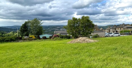 A panoramic landscape featuring a sloping green field, residential homes, and the distant hills of the Yorkshire Dales, under a cloudy sky in Cottingley, Bingley, UK.