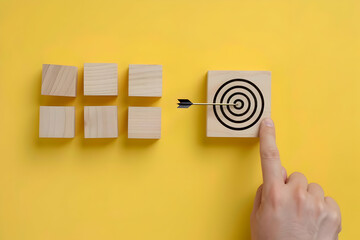 Hand setting a target on a wooden block with an arrow pointing to the center of the circle and three other blocks placed in a row above on a yellow background, representing a busin